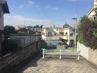 A serene residential neighborhood with a visible pathway leading up to a row of modern houses. Fences line the pathway, and lush green foliage adds color to the scene. In the background, a skyline of low-rise city buildings can be seen under a clear blue sky.