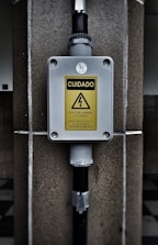 A warning sign with the text 'CUIDADO RISCO DE CHOQUE ELETRICO MANTENHA DISTANCIA DE SEGURANCA SUPERIOR A 3 METROS' in a yellow triangle, indicating an electrical shock hazard. The sign is mounted on a gray box fixed to a stone wall, with black pipes connected to the box.