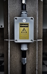 A warning sign with the text 'CUIDADO RISCO DE CHOQUE ELETRICO MANTENHA DISTANCIA DE SEGURANCA SUPERIOR A 3 METROS' in a yellow triangle, indicating an electrical shock hazard. The sign is mounted on a gray box fixed to a stone wall, with black pipes connected to the box.