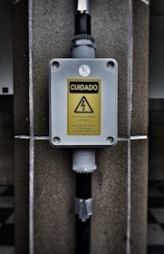 A warning sign with the text 'CUIDADO RISCO DE CHOQUE ELETRICO MANTENHA DISTANCIA DE SEGURANCA SUPERIOR A 3 METROS' in a yellow triangle, indicating an electrical shock hazard. The sign is mounted on a gray box fixed to a stone wall, with black pipes connected to the box.