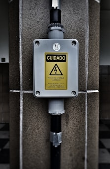 A warning sign with the text 'CUIDADO RISCO DE CHOQUE ELETRICO MANTENHA DISTANCIA DE SEGURANCA SUPERIOR A 3 METROS' in a yellow triangle, indicating an electrical shock hazard. The sign is mounted on a gray box fixed to a stone wall, with black pipes connected to the box.
