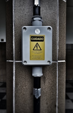 A warning sign with the text 'CUIDADO RISCO DE CHOQUE ELETRICO MANTENHA DISTANCIA DE SEGURANCA SUPERIOR A 3 METROS' in a yellow triangle, indicating an electrical shock hazard. The sign is mounted on a gray box fixed to a stone wall, with black pipes connected to the box.