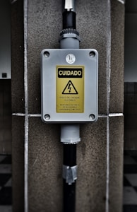 A warning sign with the text 'CUIDADO RISCO DE CHOQUE ELETRICO MANTENHA DISTANCIA DE SEGURANCA SUPERIOR A 3 METROS' in a yellow triangle, indicating an electrical shock hazard. The sign is mounted on a gray box fixed to a stone wall, with black pipes connected to the box.