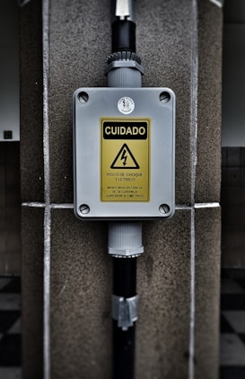 A warning sign with the text 'CUIDADO RISCO DE CHOQUE ELETRICO MANTENHA DISTANCIA DE SEGURANCA SUPERIOR A 3 METROS' in a yellow triangle, indicating an electrical shock hazard. The sign is mounted on a gray box fixed to a stone wall, with black pipes connected to the box.