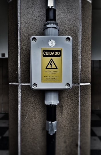 A warning sign with the text 'CUIDADO RISCO DE CHOQUE ELETRICO MANTENHA DISTANCIA DE SEGURANCA SUPERIOR A 3 METROS' in a yellow triangle, indicating an electrical shock hazard. The sign is mounted on a gray box fixed to a stone wall, with black pipes connected to the box.