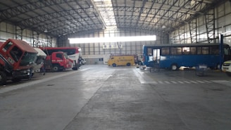 A large indoor garage or depot with a high, steel-beamed ceiling. The space contains multiple vehicles, including a red truck, a yellow armored van, and a blue bus. The vehicles seem to be parked or possibly undergoing maintenance.