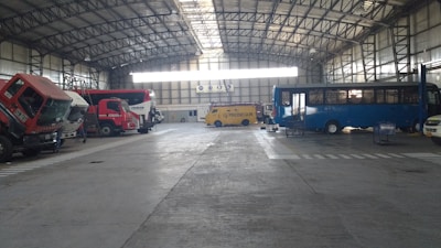 A large indoor garage or depot with a high, steel-beamed ceiling. The space contains multiple vehicles, including a red truck, a yellow armored van, and a blue bus. The vehicles seem to be parked or possibly undergoing maintenance.