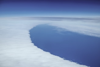 An aerial view captures a vast expanse of clouds with a clear blue sky above and a visible section of the ocean below. The scene has a serene and expansive quality, with the curvature of the earth subtly suggested.
