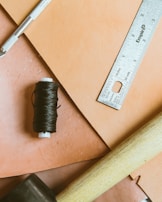 An arrangement of tools and materials including a metal ruler, a craft knife, a spool of black thread, a wooden mallet, and beige crafting sheets, set against a peach-colored background.