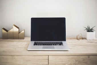 A minimalist workspace features an open laptop on a light wooden desk, flanked by a small geometric sculpture and a small potted plant. In the background, a decorative gold geometric piece adds a touch of elegance.