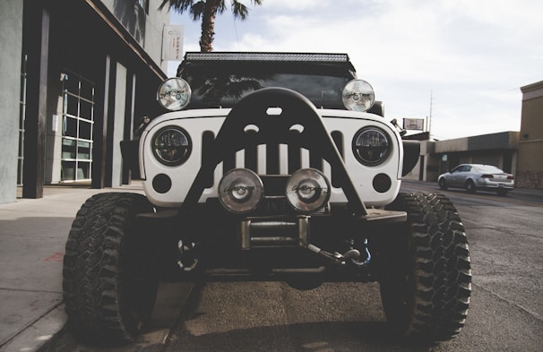 A rugged white off-road vehicle with oversized tires and additional lights is parked on an urban street. The vehicle is equipped with a front bumper guard and is positioned directly in front of the camera, creating a strong presence.