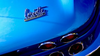 The image features a close-up of a blue Corvette Stingray, highlighting the car's rear emblem and distinct tail lights with chrome details.