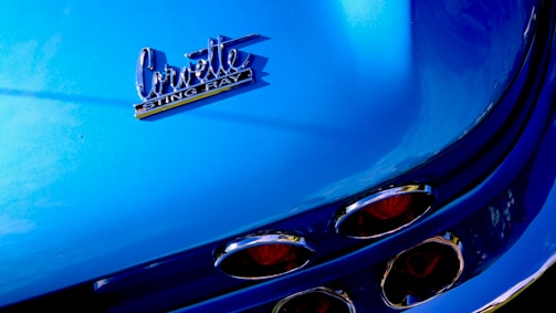 The image features a close-up of a blue Corvette Stingray, highlighting the car's rear emblem and distinct tail lights with chrome details.