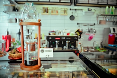 A cozy coffee shop setting featuring a cold brew coffee maker prominently displayed on a countertop. The backdrop includes a modern espresso machine, various kitchen implements, and a tiled wall with hanging utensils. Small decorative items like a plant and a colorful cloth add a touch of warmth to the minimalist decor.