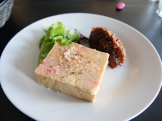 A white plate displaying a block of pâté, garnished with coarse salt and ground pepper, accompanied by a small salad of mixed greens and a portion of reddish-brown chutney or relish.