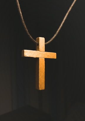 A wooden cross pendant is suspended on a dark cord against a dark background. The cross is illuminated with a warm light that highlights its texture and grain.
