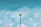 A lone security camera mounted on a tall pole with a clear blue sky and scattered clouds in the background. A bird is seen flying near the top.