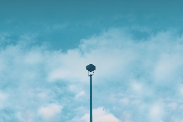 A lone security camera mounted on a tall pole with a clear blue sky and scattered clouds in the background. A bird is seen flying near the top.