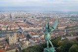Aerial view of a city featuring a large, green, winged statue in the foreground overlooking a sprawling urban landscape. The city displays a mix of historical and modern architecture with red-roofed buildings, a river running through the center, and a prominent cathedral. Trees and green spaces are interspersed amongst the buildings, adding a touch of nature to the urban setting.