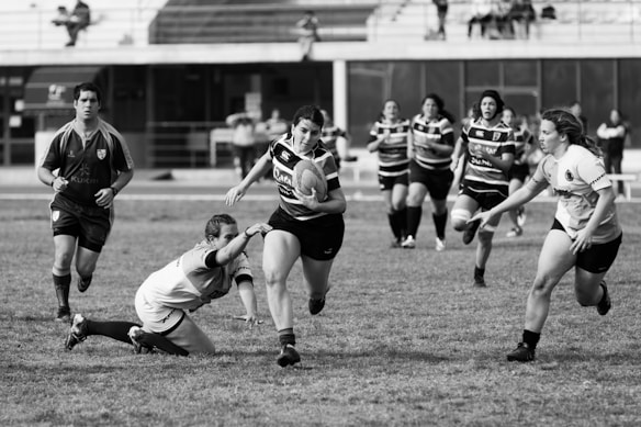 A group of women engaged in a dynamic rugby match on a field. One player is holding the ball and running towards the left, evading another player's attempt to tackle her. Other players are actively participating, showing a mixture of determination and focus. The background features empty bleachers and a partially visible referee.