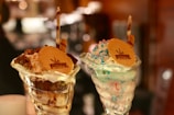Two ice cream sundaes served in elegant glass cups, topped with wafer cookies embossed with a logo. One sundae is garnished with chocolate sauce, nuts, and a chocolate stick, while the other features multi-colored sprinkles and a chocolate stick.