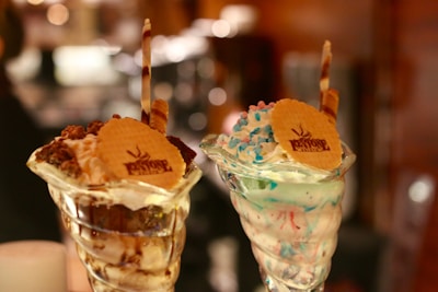 Two ice cream sundaes served in elegant glass cups, topped with wafer cookies embossed with a logo. One sundae is garnished with chocolate sauce, nuts, and a chocolate stick, while the other features multi-colored sprinkles and a chocolate stick.