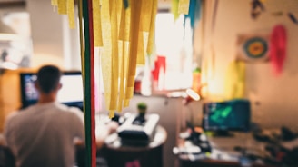 A relaxed workspace features a person sitting at a desk, with a computer screen illuminated in the foreground. Colorful paper decorations hang prominently, contributing to a creative and casual atmosphere. The decorations cast warm, cozy tones across the room, reflecting light from a small lamp on the desk.