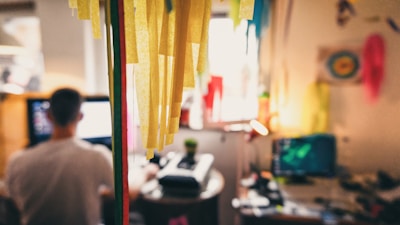 A relaxed workspace features a person sitting at a desk, with a computer screen illuminated in the foreground. Colorful paper decorations hang prominently, contributing to a creative and casual atmosphere. The decorations cast warm, cozy tones across the room, reflecting light from a small lamp on the desk.