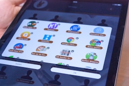 A tablet screen displaying a grid of educational and game app icons with labels, including Google Chrome, Kahoot!, and others. The screen is held in a person's hand.