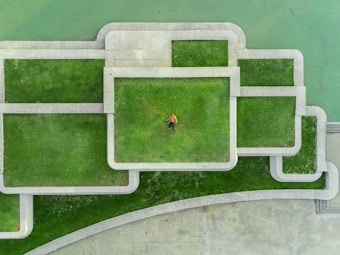 Aerial view of a geometric arrangement of green grass and concrete paths. A person wearing a yellow shirt lies at the center, providing a focal point within the structured landscape design.