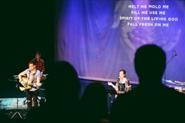 A stage setting with musicians performing under soft spotlight. Two musicians are prominent, one playing an acoustic guitar and another standing at a microphone, singing. A third person is seen in the background at a keyboard. The backdrop displays lyrics of a religious song, indicating a worship service. Audience silhouettes are visible in the foreground.