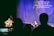 A stage setting with musicians performing under soft spotlight. Two musicians are prominent, one playing an acoustic guitar and another standing at a microphone, singing. A third person is seen in the background at a keyboard. The backdrop displays lyrics of a religious song, indicating a worship service. Audience silhouettes are visible in the foreground.