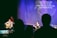A stage setting with musicians performing under soft spotlight. Two musicians are prominent, one playing an acoustic guitar and another standing at a microphone, singing. A third person is seen in the background at a keyboard. The backdrop displays lyrics of a religious song, indicating a worship service. Audience silhouettes are visible in the foreground.