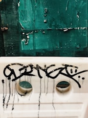 A textured green surface with peeling paint set behind a white barrier. The white barrier is marked with black graffiti, featuring drips that flow downward from the letters.