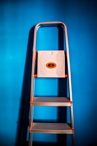 A metallic ladder with orange accents is positioned against a vibrant blue background. The ladder has wide steps and a platform at the top.