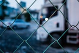 A chain-link fence in the foreground, with a blurred background showing a white truck and a car on a road or parking area.