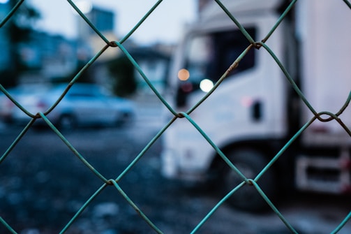 A chain-link fence in the foreground, with a blurred background showing a white truck and a car on a road or parking area.