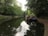 Two narrowboats are moored along the side of a calm canal, surrounded by lush, dense greenery. The boats are reflected in the still water, creating a picturesque and serene scene. A pathway lines the canal, suggesting a peaceful walkway.