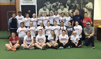 A group of women, likely a sports team, poses indoors wearing matching softball shirts with 'Lady Hawks Softball' printed on them. Most of them are smiling, and they are surrounded by a few adults, possibly coaches. In the background, there is a large black and white mural depicting historical baseball players.