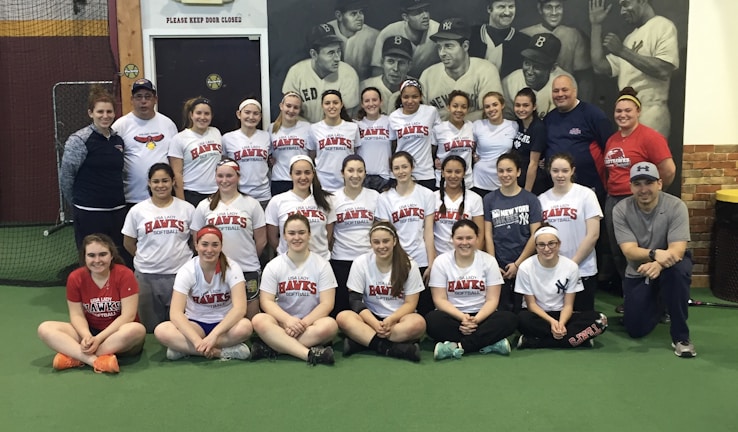 A group of women, likely a sports team, poses indoors wearing matching softball shirts with 'Lady Hawks Softball' printed on them. Most of them are smiling, and they are surrounded by a few adults, possibly coaches. In the background, there is a large black and white mural depicting historical baseball players.