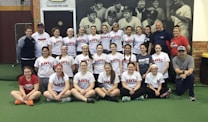 A group of women, likely a sports team, poses indoors wearing matching softball shirts with 'Lady Hawks Softball' printed on them. Most of them are smiling, and they are surrounded by a few adults, possibly coaches. In the background, there is a large black and white mural depicting historical baseball players.