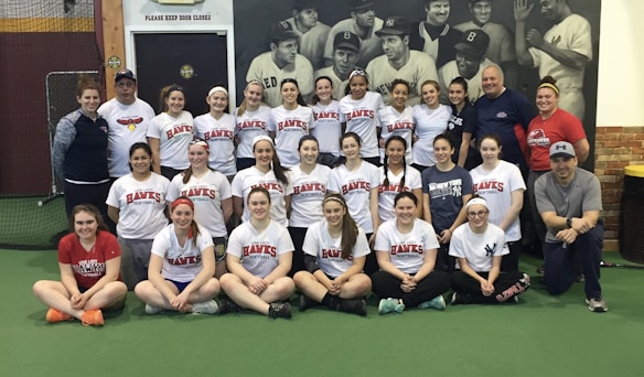 A group of women, likely a sports team, poses indoors wearing matching softball shirts with 'Lady Hawks Softball' printed on them. Most of them are smiling, and they are surrounded by a few adults, possibly coaches. In the background, there is a large black and white mural depicting historical baseball players.