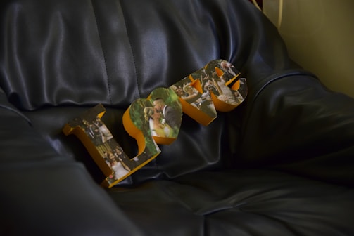 Wooden letters spelling 'LOVE' resting on a dark leather couch, with each letter featuring collage-style photographs of wedding scenes, including a bride and groom.