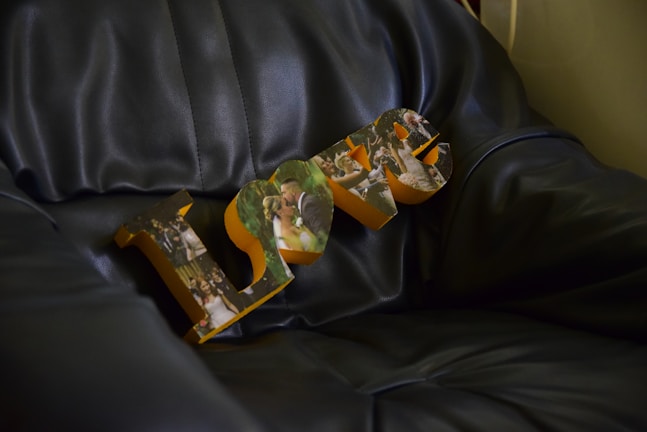 Wooden letters spelling 'LOVE' resting on a dark leather couch, with each letter featuring collage-style photographs of wedding scenes, including a bride and groom.