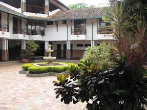 A courtyard with a central stone fountain surrounded by neatly trimmed green bushes. The area is paved with red and brown bricks. The background features a two-story building with a tiled roof and white walls, accented by dark green columns and window frames. Several potted plants add to the lush greenery, and there is a variety of foliage in the foreground.