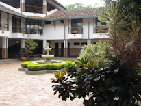 A courtyard with a central stone fountain surrounded by neatly trimmed green bushes. The area is paved with red and brown bricks. The background features a two-story building with a tiled roof and white walls, accented by dark green columns and window frames. Several potted plants add to the lush greenery, and there is a variety of foliage in the foreground.