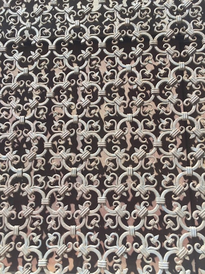 Intricate wrought iron pattern featuring repeating loops and floral motifs, creating a complex and artistic lattice. The metal appears weathered, adding a vintage or antique feel.