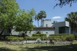 A historic building with white stucco walls and a red-tiled roof is surrounded by lush greenery and palm trees. In front of the building, there is a small decorative pond surrounded by manicured shrubs and a well-maintained lawn. The sky is clear with a few scattered clouds, creating a serene ambiance.