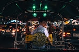An interior view of a tuk-tuk taxi navigating through busy nighttime traffic. The driver, wearing a light blue shirt, is looking back toward the camera. Bright lights from surrounding cars illuminate the scene, reflecting off various surfaces.
