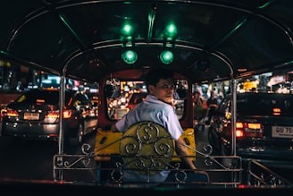 An interior view of a tuk-tuk taxi navigating through busy nighttime traffic. The driver, wearing a light blue shirt, is looking back toward the camera. Bright lights from surrounding cars illuminate the scene, reflecting off various surfaces.